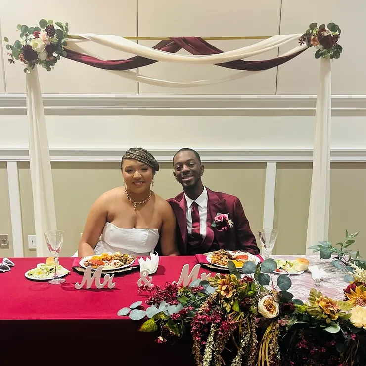Mr and Mrs at burgundy sweetheart table with draped arch and autumn florals