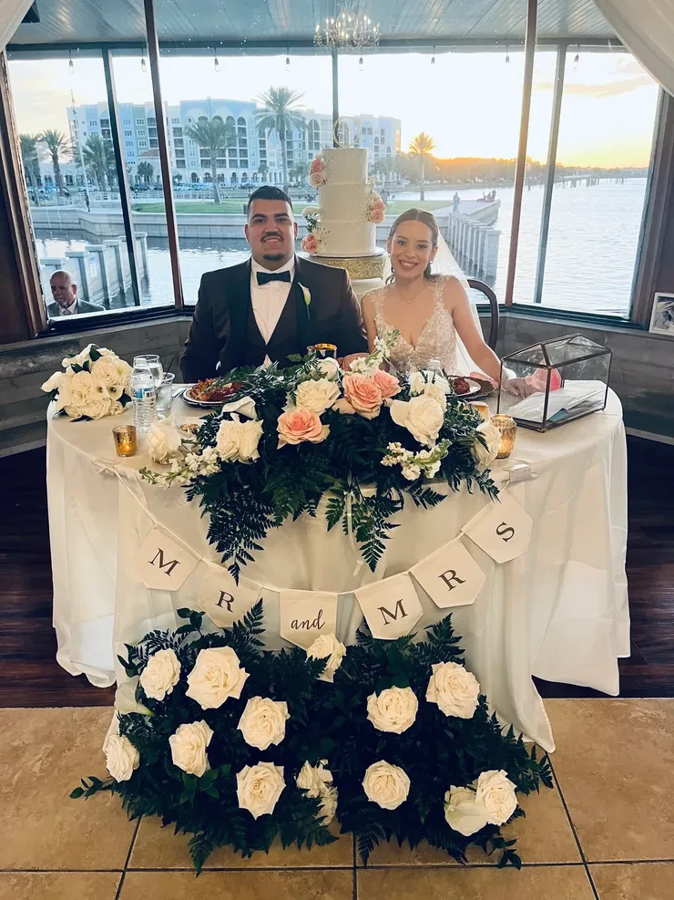 Mr and Mrs at waterfront venue sweetheart table with white roses and wedding cake backdrop